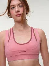 Top deportivo rosa con efecto layering, escote redondo bajo y tirantes dobles. Presenta el logo de la marca en tono oscuro en el frente.