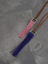 Collar con cordón de cuero marrón y dije cuadrado metálico plateado del cual cuelga una borla de hilos de seda en color azul.