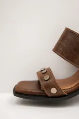 Sandalias de cuero marrón, con taco ancho, faja ancha en el empeine, tira frontal con tachas metálicas y punta cuadrada. Cuentan con ajuste mediante hebilla al tobillo.