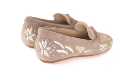 Mocasín de gamuza color avena con bordados florales en hilo blanco y pequeños apliques metálicos dorados. Presenta un lazo decorativo en la capellada y suela de goma flexible.