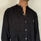 Camisa de lino blanca con cuello mao y cierre frontal de botones. Presenta un pequeño bordado oscuro en el pecho y mangas largas con puños doblados.