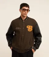 Campera bomber de estilo college con cuerpo a cuadros en tonos marrones y mangas largas de cuero sintético negro. Presenta cuello, puños y cintura acanalados a rayas, cierre abotonado, bolsillos laterales y aplique bordado de letra "B" en el pecho.