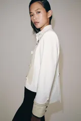 Chaqueta de estilo militar de ZARA Woman Collection, confeccionada en mezcla de algodón en color blanco y beige. Presenta cuello subido, manga larga con puños anchos en contraste y cierre frontal con alamares y botones metálicos en relieve. Tiene bajo asimétrico con abertura en la espalda.