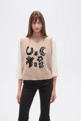 Chaleco de punto sin mangas en color beige con efecto confetti y diseño de jacquard frontal en color negro con motivos de herradura, luna y pájaro. Cuenta con cuello redondo y acabados acanalados.
