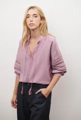 Camisa de color crudo con diseño romántico, cuello redondo con cordón ajustable terminado en borlas, y mangas tres cuartos abullonadas. Presenta detalles de bordados calados verticales en el frente y horizontales en las mangas.