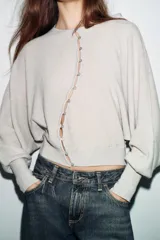 Chaqueta de punto color beige claro, de cuello redondo y manga larga abullonada con puño ajustado. Cierre frontal cruzado con botones plateados.