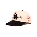 Gorra con visera plana de color negro y corona en tono natural. Presenta bordados frontales en color negro con el monograma central y las frases "Mi Casa" y "Su Casa" a los lados.