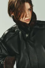 Cazadora bomber de efecto piel en color negro, con cuello subido, manga larga y bajo elástico. Presenta interior y puños con aplicación de pelo sintético en color marrón a contraste, cierre frontal con cremallera y botones a presión, y bolsillos laterales de vivo.