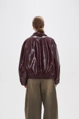 Chaqueta de eco-cuero en color bordó con acabado brillante, diseño holgado, cuello camisero, cierre frontal con botones a presión y detalle de elástico fruncido en el ruedo.