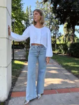 Blusa blanca de algodón con textura arrugada, cuello redondo, mangas largas abullonadas con puños elásticos y cierre frontal con botones y volantes.