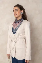 Chaquetón color beige de corte recto y largo, con cierre frontal mediante botones y lazo ajustable en la cintura. Presenta cuello en V, bolsillos laterales con solapa y mangas largas.