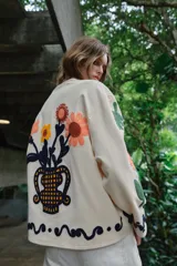 Chaqueta de algodón color crudo con cuello redondo, manga larga y bolsillos delanteros de plastrón. Presenta un diseño con bordados coloridos de flores y motivos abstractos a contraste, y cierre frontal con botones oscuros.