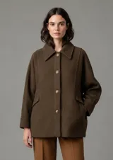 Abrigo de paño color marrón oscuro, con cuello camisero, cierre frontal de botones metálicos y bolsillos laterales con solapa. Presenta un corte holgado y mangas largas.