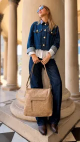 Cartera de tamaño mediano confeccionada en símil cuero color tostado, con diseño de bolsillo frontal con tapa y cierre, correa larga regulable y asa de mano.