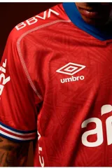 Camiseta de fútbol Umbro del Club Nacional de Football, modelo Away 2025 de color rojo con detalles en azul en el cuello en V y en las mangas. Cuenta con patrocinadores impresos en blanco, el logo de la marca y el escudo del club.