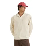 Remera de manga larga tipo polo, color beige, confeccionada en tejido de textura tipo toalla (terry). Presenta cuello camisero, cierre frontal con botones ocultos y un bolsillo aplicado en el pecho.