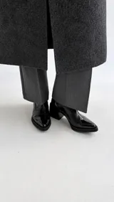 Botinetas de cuero negro con acabado brillante, punta fina y taco cuadrado bajo. Presentan un diseño minimalista con costuras visibles en la puntera y cierre lateral.