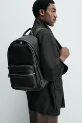 Mochila de diseño minimalista en color negro, confeccionada en material sintético con acabado grabado. Cuenta con un compartimento principal con cierre de cremallera, un bolsillo frontal con cierre y un compartimento posterior acolchado para dispositivos electrónicos. Incluye asa de mano superior y dos correas de hombro ajustables.