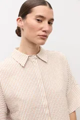Camisa de manga corta con diseño a rayas en tonos beige y blanco, cuello camisero y cierre frontal con botones ocultos.