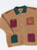 Campera tejida de punto grueso con diseño multicolor. Presenta cuello en contraste de color verde, cuerpo en tono beige con textura trenzada, bolsillos delanteros con apliques en nylon color marrón y bordó, y detalles en nylon en los hombros. Cuenta con cierre frontal de botones y forro interno.