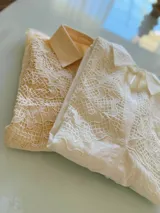 Camisa de manga larga en color beige, con cuello clásico y cierre frontal de botones. Presenta un delicado aplique de encaje tipo crunie en la parte superior delantera, aportando textura y un detalle artesanal al diseño.