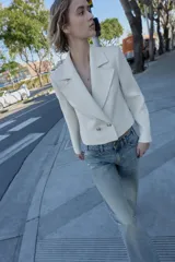 Blazer corto blanco de cuello solapa y manga larga con hombreras. Presenta un cierre frontal cruzado con dos botones dorados.