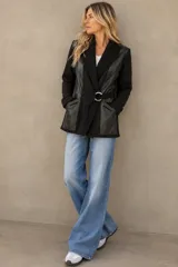Chaqueta negra combinada de lana merino y cuero sintético, con cuello tipo chal tejido, bolsillos frontales y cinto con hebilla metálica circular.