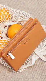 Billetera de mujer tipo sobre con correa de mano extraíble, confeccionada en material texturizado color tostado. Presenta un bolsillo frontal con cierre, compartimentos internos para tarjetas y logo de la marca grabado en el frente.