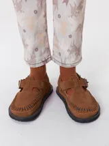 Slipper tipo guillermina confeccionada en gamuza color marrón, con plataforma de goma negra y doble hebilla ajustable. Presenta un pespunte decorativo en el borde inferior y cuenta con interior recubierto en cuero.