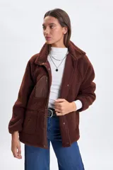 Campera de sherpa color marrón con cuello alto, cierre frontal con botones a presión y detalles en tela técnica a tono. Presenta un bolsillo vertical con cierre en el pecho y dos bolsillos laterales con cierre.
