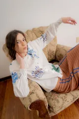 Camisa de popelina de algodón color crudo, con cuello bobo amplio con volados y cierre frontal de botones. Presenta un diseño estampado con motivos botánicos y de aves en tonos azules y verdes.