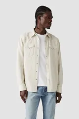 Camisa de manga larga color beige, con cierre de botones a presión, dos bolsillos frontales con solapa y logo de la marca en etiqueta roja en el bolsillo izquierdo.
