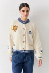 Cardigan de lana color crema con cuello redondo, cierre frontal de botones y mangas largas con puños ajustados. Presenta bordados decorativos en el frente que incluyen un sol, olas y estrellas, con detalles en azul en el cuello y el ruedo.
