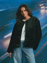 Campera de textura afelpada en color negro, con cuello redondo, cierre frontal con botones y bolsillos delanteros. Presenta detalles de terminaciones en material sintético liso a lo largo de la abertura y los bolsillos.