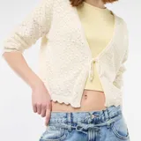 Chaqueta de punto con textura de croché en color beige, de corte corto y mangas largas. Presenta un cierre frontal mediante una lazada fina y bordes con acabado festoneado.