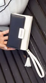 Billetera mediana de diseño bicolor, combinando una solapa en tono dorado metalizado con cuerpo en color negro. Presenta cierre perimetral, correa de muñeca extraíble con detalle de borla y placa metálica con logo de la marca en el frente.