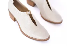 Zapato cerrado tipo mocasín con taco cuadrado de madera, confeccionado en cuero con textura de reptil en color crema. Presenta un detalle de hebilla metálica dorada en el empeine.