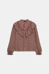 Blusa de manga larga con estampado de cuadros vichy en tonos burdeos y beige. Presenta cuello mao y grandes volantes fruncidos que cruzan el pecho y los hombros. Los puños son elásticos y fruncidos.