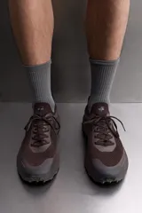 Zapatilla deportiva de correr para trail running, color marrón oscuro. Presenta una parte superior de tejido de punto transpirable, puntera y talón reforzados con material Shield Protect Max. Cuenta con ajuste de cordones, tirador trasero, plantilla OrthoLite® extraíble, entresuela Spume Foam V2 y suela de goma.