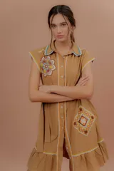 Vestido corto beige de algodón con cuello camisero y cierre frontal de botones. Presenta mangas cortas y detalles bordados a contratono en la zona del pecho y en los bolsillos, además de un volado en el ruedo.