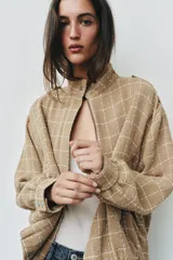 Chaqueta bomber de color beige con diseño a cuadros blancos, cuello subido, manga larga con trabillas en los hombros, bolsillos delanteros y bajo con elástico. Cierre frontal con cremallera y botones a presión.
