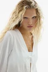 Blusa blanca de algodón con escote en V, frente abierto con cierre de tiras finas para anudar, mangas largas y puños elásticos.
