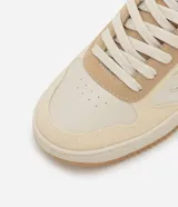 Championes casuales de caña baja, color beige y crema, con suela tipo caja. Presentan una combinación de materiales lisos y gamuza sintética, perforaciones en la punta para ventilación, cierre de cordones y punta redondeada.