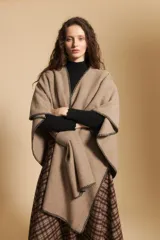 Poncho de tejido suave color beige con diseño envolvente y terminaciones con costuras decorativas en contraste color negro. Presenta un detalle de lazo frontal integrado.