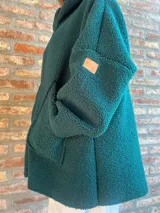Campera de corderito color verde oscuro, con diseño holgado, cuello alto y manga larga. Presenta una etiqueta de cuero sintético en la manga.