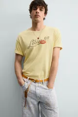 Camiseta de corte relajado color amarillo con efecto lavado. Presenta cuello redondo, mangas cortas con dobladillo y un estampado frontal con texto y un parche decorativo.
