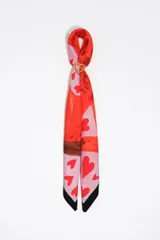 Pañuelo rectangular de poliéster con estampado artístico en tonos rojos, rosados y negros, que incluye un diseño de corazones. Viene acompañado de un charm metálico dorado en forma de corazón.