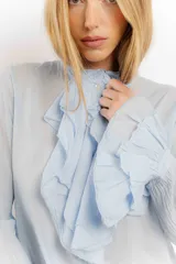 Camisa de estilo romántico confeccionada en algodón, con cuello alto, volados frontales y mangas largas con puños acampanados también decorados con volados.