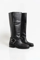 Botas de caña alta confeccionadas en cuero negro, con diseño de estilo western que incluye una correa decorativa con argollas metálicas y remaches en el tobillo. Presentan una puntera cuadrada y un taco bajo cuadrado.