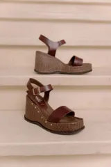 Sandalias de plataforma y taco alto confeccionadas en cuero vacuno marrón, con tira ancha en el empeine, tira ajustable con hebilla en el tobillo y detalles de tachas decorativas en la base.